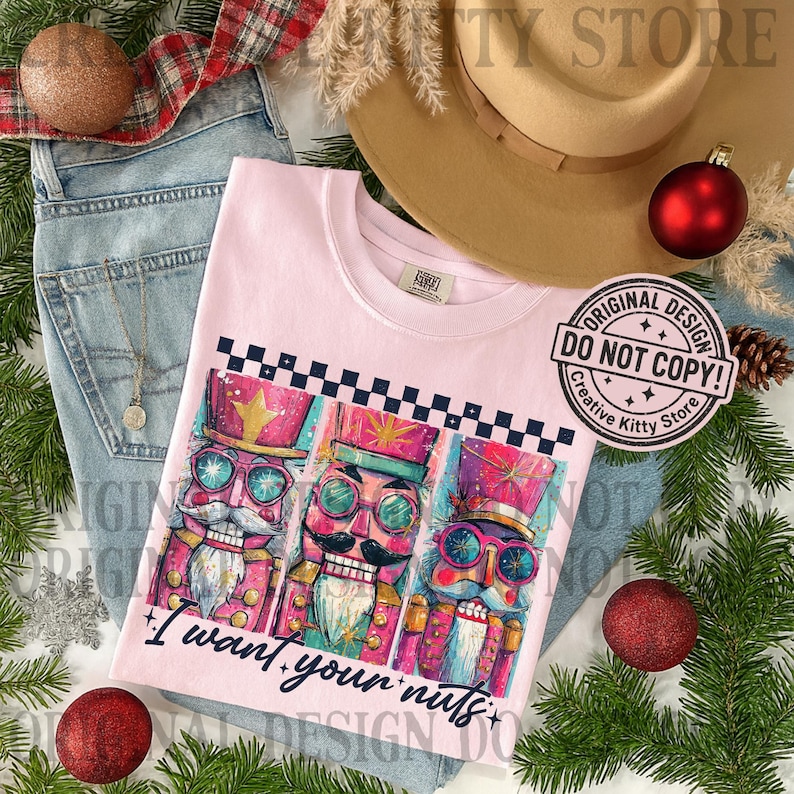 May include: A light pink t-shirt featuring a graphic of three colorful nutcrackers with sunglasses and the text "I want your nuts." The shirt is displayed with blue jeans, a hat, and Christmas ornaments.