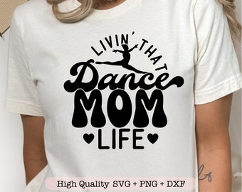 May include: Cream-colored t-shirt with a black graphic that reads "LIVIN' THAT Dance MOM LIFE" with a silhouette of a dancer. Two black hearts flank the word "LIFE". The text "High Quality SVG + PNG + DXF" is at the bottom.