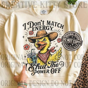 May include: Cream-colored sweatshirt with a graphic of a yellow duck wearing a cowboy hat and bandana, holding a drink. Text reads "I Don't Match Energy" and "Shut The Power Off". Features a "Do Not Copy" design stamp.