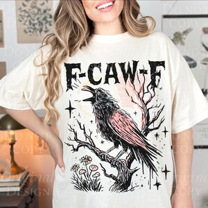 May include: Cream-colored t-shirt featuring a graphic of a crow perched on a branch, with the text "F-CAW-F". The design includes a black crow with pink accents, bare tree branches, and stars. The shirt has a relaxed fit.