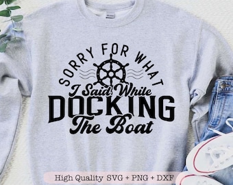 May include: A gray sweatshirt with the text "Sorry for what I said while docking the boat" in black. A black and white illustration of a ship's wheel is above the text. The text "High Quality SVG + PNG + DXF" is at the bottom of the image.