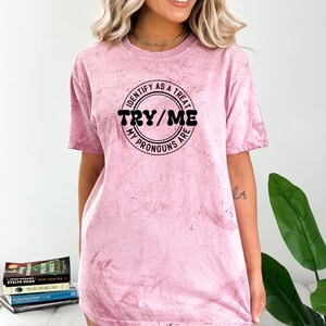 Puede incluir: Una camiseta rosa tie-dye con un gr&aacute;fico circular negro que dice "Identify as a treat, Try/Me, My pronouns are".