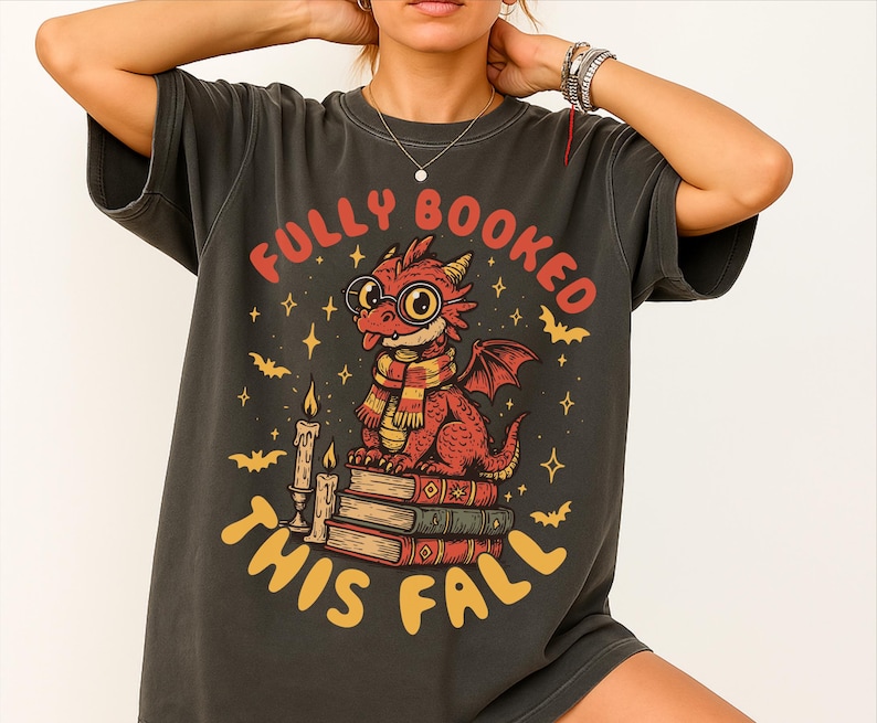 K&ouml;nnte beinhalten: Dunkelgraues T-Shirt mit einem Motiv eines roten Drachen mit Brille und Schal, der auf B&uuml;chern sitzt. Der Text "FULLY BOOKED THIS FALL" ist in Rot und Gelb gehalten. Flederm&auml;use, Sterne und Kerzen erg&auml;nzen das Design.