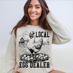Op de afbeelding: Een wit sweatshirt met een zwart-witte illustratie van een kip met eieren en de tekst "Local Egg Dealer".