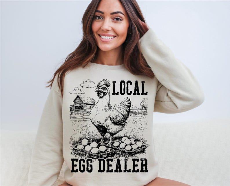 Op de afbeelding: Een wit sweatshirt met een zwart-witte illustratie van een kip met eieren en de tekst "Local Egg Dealer".