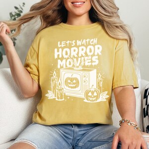 May include: A yellow t-shirt with a white graphic of a vintage television with a pumpkin on the screen. The text "Let's Watch Horror Movies" is above the television. There are candles and pumpkins on either side of the television.