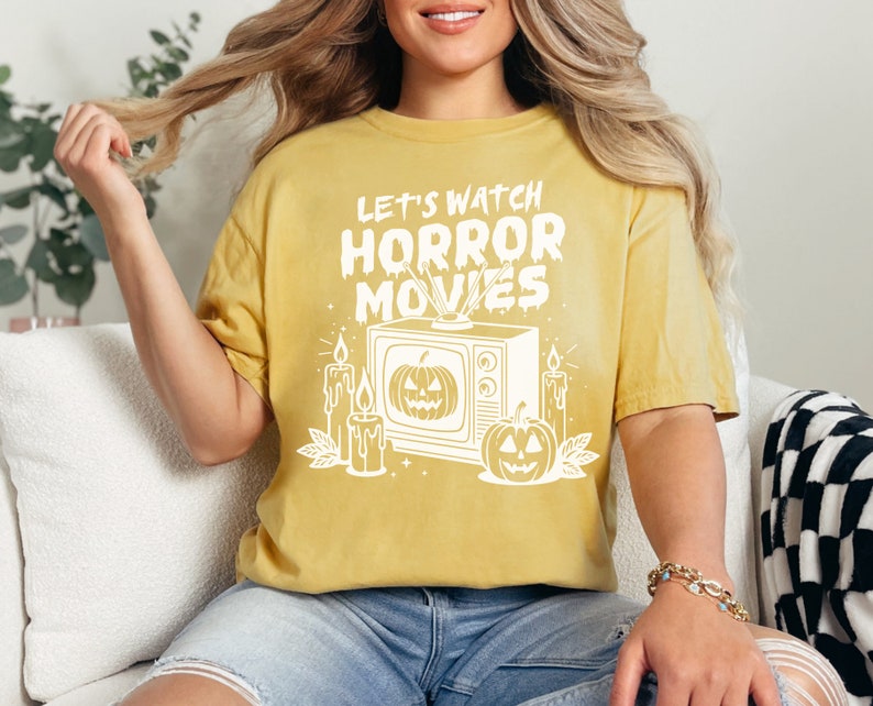 May include: A yellow t-shirt with a white graphic of a vintage television with a pumpkin on the screen. The text "Let's Watch Horror Movies" is above the television. There are candles and pumpkins on either side of the television.