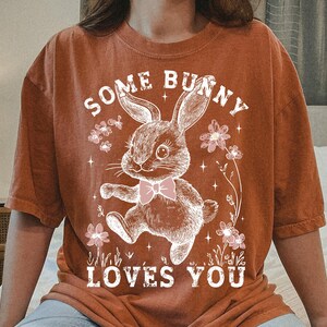 Peut inclure: Un t-shirt couleur rouille avec un graphique de lapin blanc et le texte "Some Bunny Loves You".