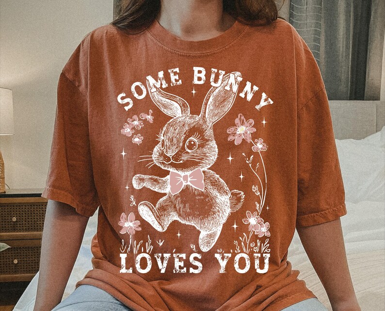 Peut inclure: Un t-shirt couleur rouille avec un graphique de lapin blanc et le texte "Some Bunny Loves You".