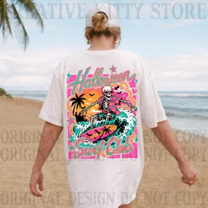 May include: A white t-shirt featuring a colorful graphic on the back. The design shows a skeleton wearing sunglasses and a floral shirt, surfing on a wave. The text reads Halloween Surf Club, surrounded by palm trees, bats, and stars.