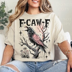 May include: Beige t-shirt with a graphic of a crow perched on a branch, with the text "F-CAW-F". The crow is black with pink accents. The design includes stars and flowers.