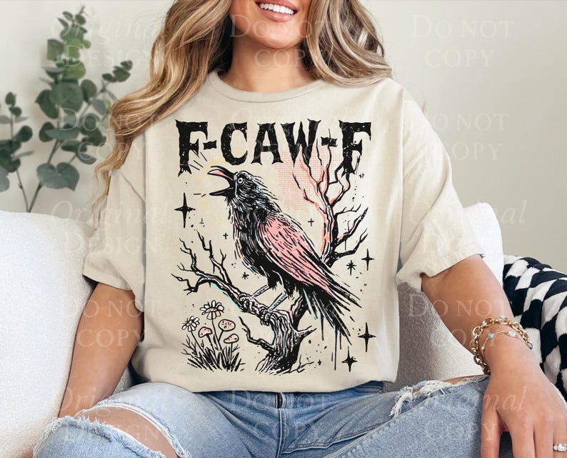 May include: Beige t-shirt with a graphic of a crow perched on a branch, with the text "F-CAW-F". The crow is black with pink accents. The design includes stars and flowers.