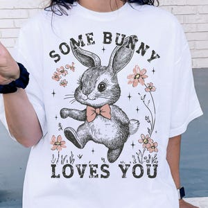 Peut inclure: T-shirt blanc avec une illustration en noir et blanc d'un lapin avec le texte "Some Bunny Loves You" en noir.