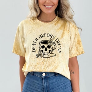 May include: A yellow tie-dye effect t-shirt with short sleeves. The black graphic on the front depicts a skull within a coffee cup, adorned with roses, and the text "DEATH BEFORE DECAF" in a circular arrangement.