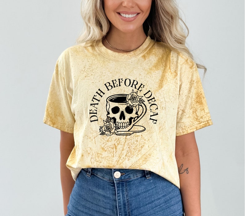 May include: A yellow tie-dye effect t-shirt with short sleeves. The black graphic on the front depicts a skull within a coffee cup, adorned with roses, and the text "DEATH BEFORE DECAF" in a circular arrangement.