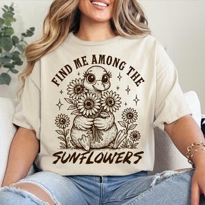 May include: A beige t-shirt with a graphic of a duck holding a bouquet of sunflowers. The text "Find me among the sunflowers" is printed on the shirt.