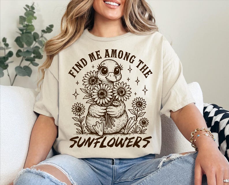 May include: A beige t-shirt with a graphic of a duck holding a bouquet of sunflowers. The text "Find me among the sunflowers" is printed on the shirt.