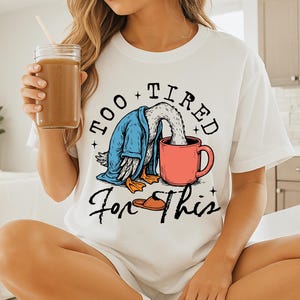 Op de afbeelding: Wit T-shirt met een afbeelding van een cartoon eend die een blauwe badjas draagt en drinkt uit een rode mok. De tekst "Too Tired For This" is op het T-shirt gedrukt.