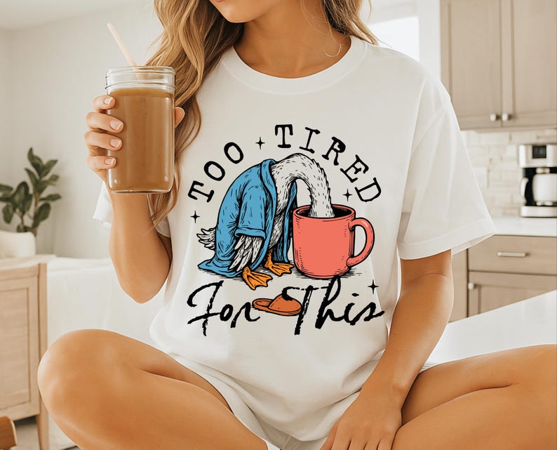 Op de afbeelding: Wit T-shirt met een afbeelding van een cartoon eend die een blauwe badjas draagt en drinkt uit een rode mok. De tekst "Too Tired For This" is op het T-shirt gedrukt.