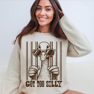 Pu&ograve; includere: Una felpa beige con una grafica di un'anatra che indossa occhiali da sole e il testo "GOT TOO SILLY" stampato sul davanti.
