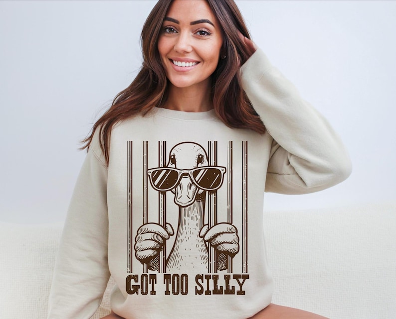 Pu&ograve; includere: Una felpa beige con una grafica di un'anatra che indossa occhiali da sole e il testo "GOT TOO SILLY" stampato sul davanti.