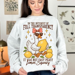 May include: A white sweatshirt with a graphic of a duck wearing sunglasses and holding a lemon. The text on the sweatshirt reads "In the interest of full transparency it was not easy peasy lemon squeezy."