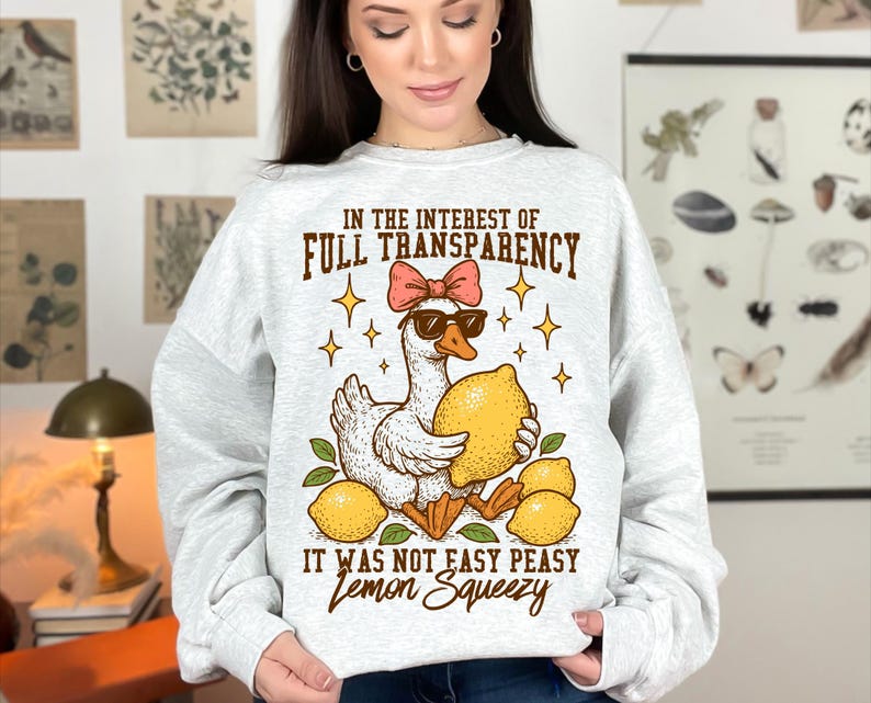 May include: A white sweatshirt with a graphic of a duck wearing sunglasses and holding a lemon. The text on the sweatshirt reads "In the interest of full transparency it was not easy peasy lemon squeezy."