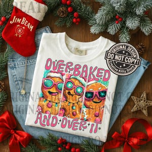 Puede incluir: Camiseta blanca con el texto "OVERBAKED AND OVER IT" y un gr&aacute;fico de tres hombres de jengibre con gafas de sol y gorros de Pap&aacute; Noel. La camiseta est&aacute; sobre unos vaqueros azules, con un calcet&iacute;n rojo y adornos navide&ntilde;os.