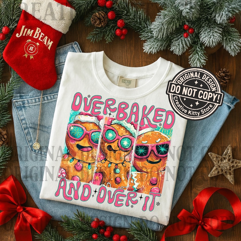 Puede incluir: Camiseta blanca con el texto "OVERBAKED AND OVER IT" y un gr&aacute;fico de tres hombres de jengibre con gafas de sol y gorros de Pap&aacute; Noel. La camiseta est&aacute; sobre unos vaqueros azules, con un calcet&iacute;n rojo y adornos navide&ntilde;os.