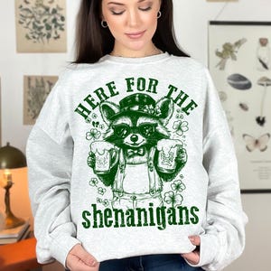 Puede incluir: Una sudadera gris con un gr&aacute;fico verde de un mapache con gafas de sol y sosteniendo dos jarras de cerveza. El texto "Here for the shenanigans" est&aacute; impreso debajo del mapache.