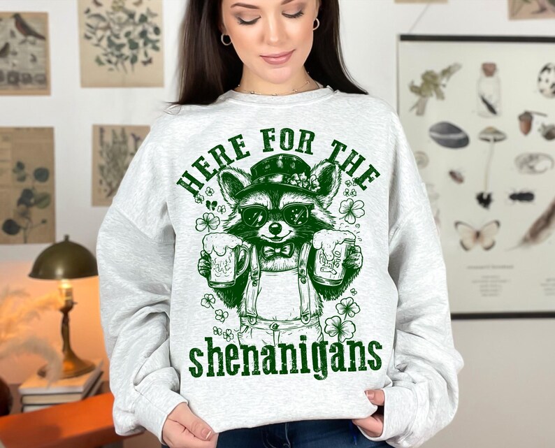 Puede incluir: Una sudadera gris con un gr&aacute;fico verde de un mapache con gafas de sol y sosteniendo dos jarras de cerveza. El texto "Here for the shenanigans" est&aacute; impreso debajo del mapache.