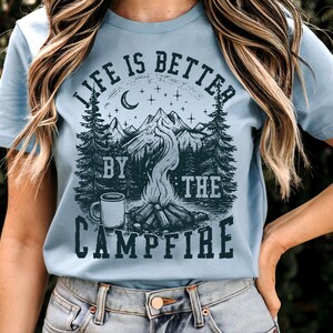 Puede incluir: Camiseta azul claro con un dise&ntilde;o gr&aacute;fico de una escena de fogata. El dise&ntilde;o incluye monta&ntilde;as, &aacute;rboles, una luna creciente, estrellas y el texto "LIFE IS BETTER BY THE CAMPFIRE" en azul oscuro. La camiseta es de material suave.