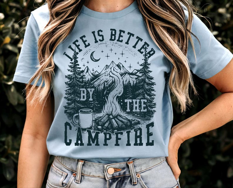 Puede incluir: Camiseta azul claro con un dise&ntilde;o gr&aacute;fico de una escena de fogata. El dise&ntilde;o incluye monta&ntilde;as, &aacute;rboles, una luna creciente, estrellas y el texto "LIFE IS BETTER BY THE CAMPFIRE" en azul oscuro. La camiseta es de material suave.