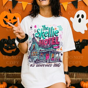 K&ouml;nnte beinhalten: Wei&szlig;es T-Shirt mit einer farbenfrohen Illustration von "The Skellie Motel" mit einem gruseligen Hausdesign, Spinnweben und dem Text "Old Graveyard Road". Das Shirt hat ein Halloween-Thema.