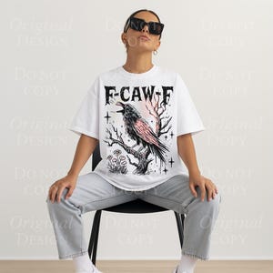 May include: White t-shirt with a graphic of a crow perched on a branch, with the text "F-CAW-F" in a gothic font. The crow is black and pink, with a pink and orange background. The shirt is worn by a person.