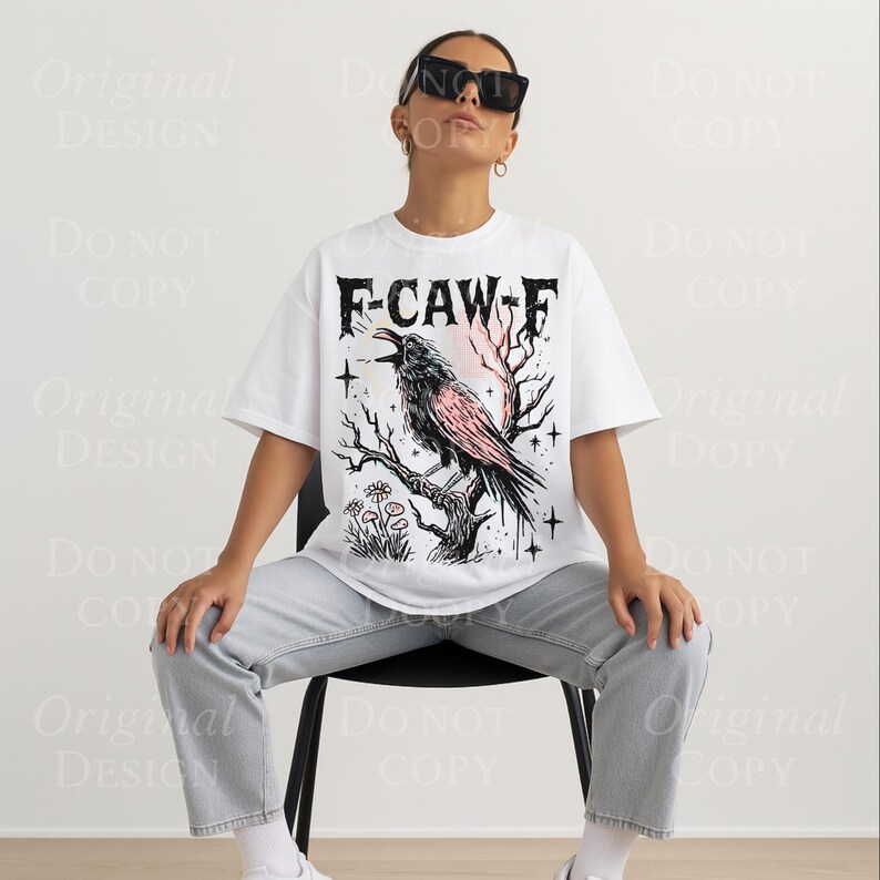 May include: White t-shirt with a graphic of a crow perched on a branch, with the text "F-CAW-F" in a gothic font. The crow is black and pink, with a pink and orange background. The shirt is worn by a person.