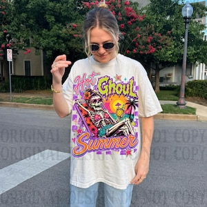 May include: An off-white short-sleeve t-shirt featuring a graphic print of a skeleton lounging in a beach chair. The text reads Hot Ghoul Summer. The design includes a sunset, palm tree, and tropical drink in vibrant shades of pink, orange, and purple.