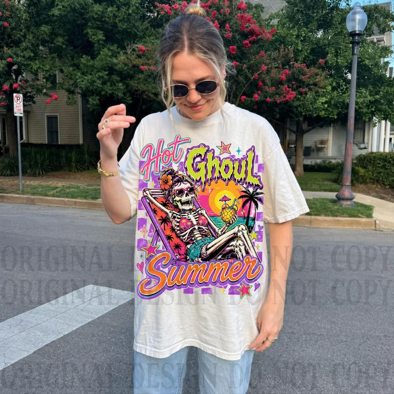 May include: An off-white short-sleeve t-shirt featuring a graphic print of a skeleton lounging in a beach chair. The text reads Hot Ghoul Summer. The design includes a sunset, palm tree, and tropical drink in vibrant shades of pink, orange, and purple.