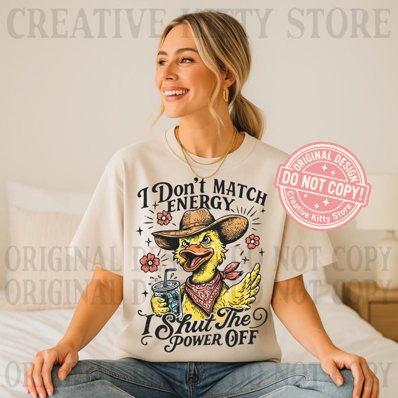 May include: Cream-colored t-shirt featuring a cartoon duck wearing a cowboy hat and bandana, holding a drink. The shirt has the text "I Don't Match Energy, I Shut The Power Off" with floral accents. A pink "Original Design" stamp is visible.
