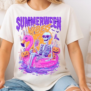 May include: A cream-coloured t-shirt with a vibrant graphic. The design shows a skeleton in sunglasses, lounging on a pink flamingo float, holding a drink, with pumpkins and candles. The text "Summerween Vibes" is displayed above the image.