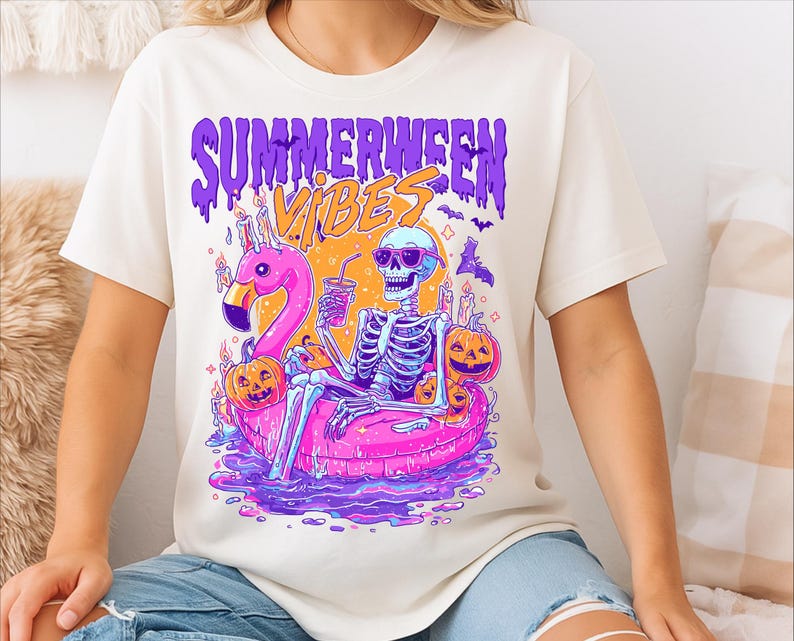 May include: A cream-coloured t-shirt with a vibrant graphic. The design shows a skeleton in sunglasses, lounging on a pink flamingo float, holding a drink, with pumpkins and candles. The text "Summerween Vibes" is displayed above the image.