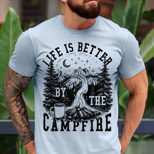 Puede incluir: Camiseta azul claro con un dise&ntilde;o gr&aacute;fico negro. El dise&ntilde;o presenta una escena de fogata con monta&ntilde;as, &aacute;rboles, una luna creciente, estrellas y el texto "LIFE IS BETTER BY THE CAMPFIRE".