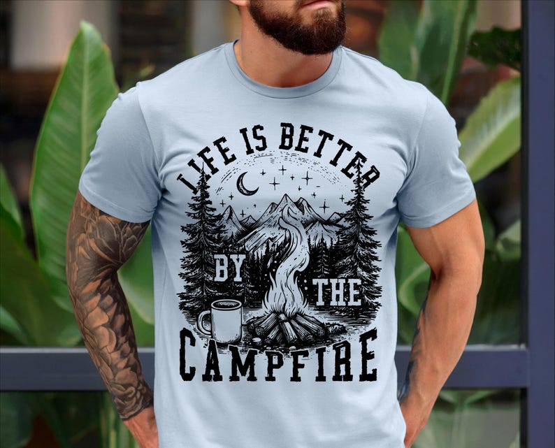 Puede incluir: Camiseta azul claro con un dise&ntilde;o gr&aacute;fico negro. El dise&ntilde;o presenta una escena de fogata con monta&ntilde;as, &aacute;rboles, una luna creciente, estrellas y el texto "LIFE IS BETTER BY THE CAMPFIRE".