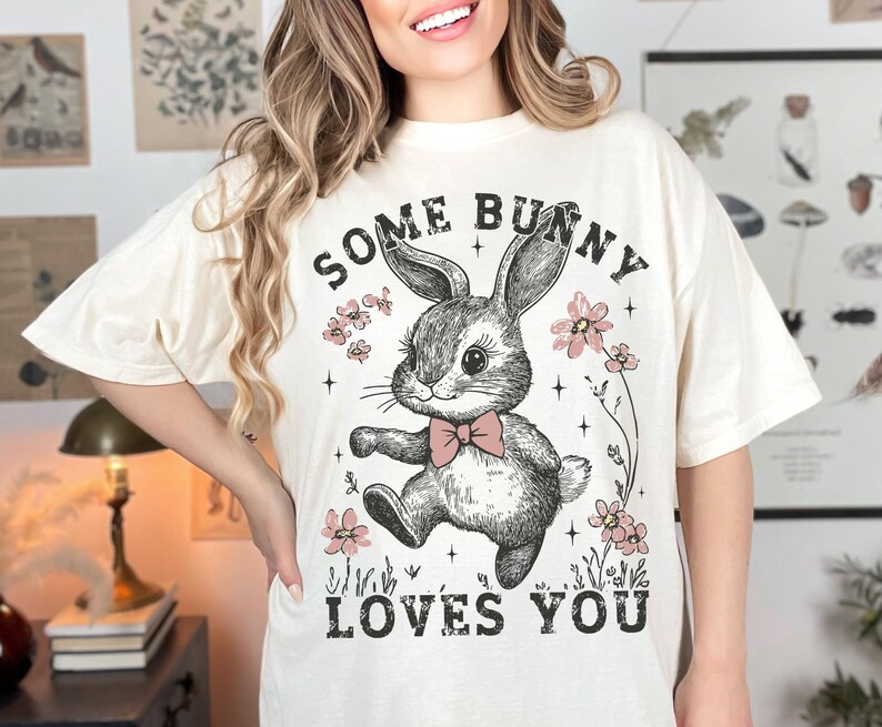 Peut inclure: Un t-shirt blanc avec un motif de lapin de dessin anim&eacute; avec des fleurs roses et le texte "Some Bunny Loves You".