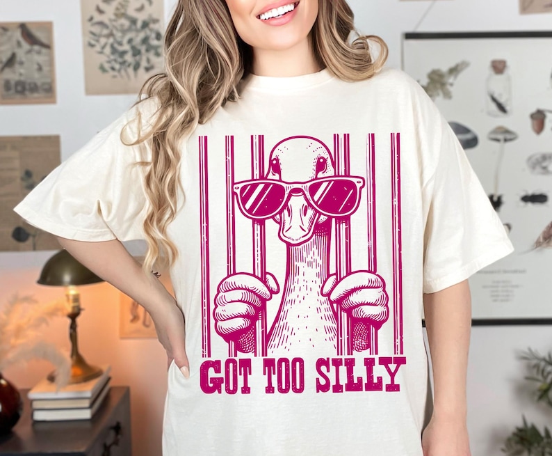 Pu&ograve; includere: Maglietta bianca con una grafica rosa di un'anatra con occhiali da sole e dietro le sbarre. Il testo "Got Too Silly" &egrave; sotto la grafica.