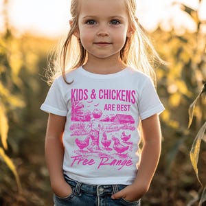K&ouml;nnte beinhalten: Ein wei&szlig;es T-Shirt mit einem pinkfarbenen Grafikdesign, das ein Kind und H&uuml;hner zeigt. Die Grafik lautet "Kids & Chickens Are Best Free Range".