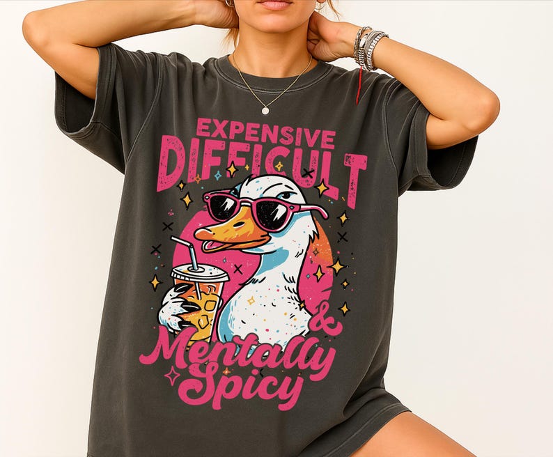 Pu&ograve; includere: T-shirt grigio scuro con un'anatra dei cartoni animati con occhiali da sole e una bevanda. La maglietta ha il testo "EXPENSIVE DIFFICULT & Mentally Spicy" in rosa e bianco. L'anatra &egrave; bianca, arancione e blu.