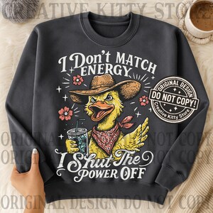 May include: Dark gray sweatshirt with a graphic of a yellow duck wearing a cowboy hat and bandana, holding a drink. Text reads "I Don't Match Energy, I Shut The Power Off." Features a "Do Not Copy" design.