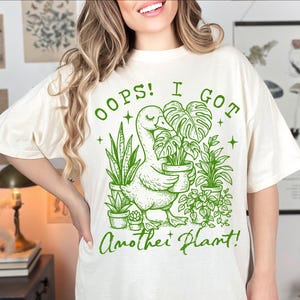 May include: A white t-shirt with a green graphic of a duck carrying a potted plant. The text on the shirt reads "Oops! I got another plant!"