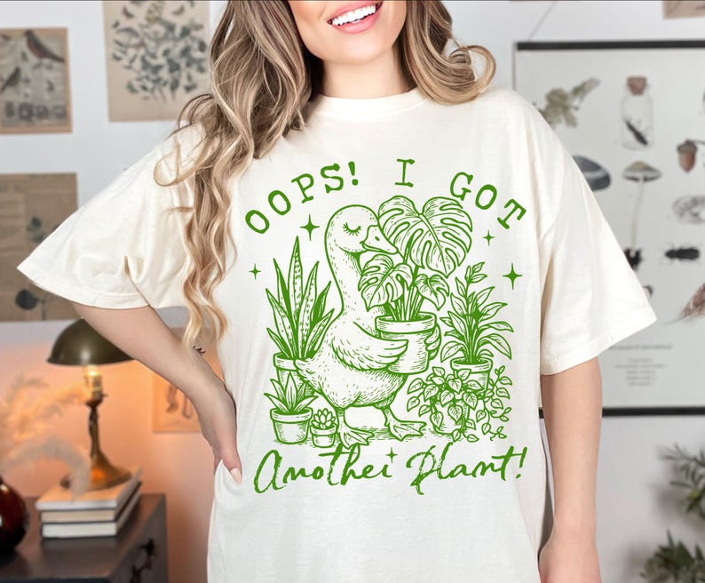 May include: A white t-shirt with a green graphic of a duck carrying a potted plant. The text on the shirt reads "Oops! I got another plant!"